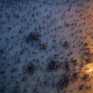 A vast crowd of people photographed from directly overhead in an open stone plaza at dusk, hundreds of small figures scattered unevenly, some clustered into dense knots and others standing alone, each casting a long soft shadow across the pale paving. Cool blue twilight with warm amber light spilling in from one edge, muted desaturated palette, fine grain, cinematic aerial composition with shallow atmospheric haze.