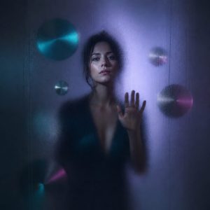 A woman in her thirties stands behind a large pane of frosted glass in a dark room, her figure clear at the centre and dissolving into blur toward the edges, lit by cool violet light from one side. Smooth metallic discs float in the dim air around her, half hidden behind the glass. Teal and magenta accents, high contrast, cinematic shallow depth of field.
